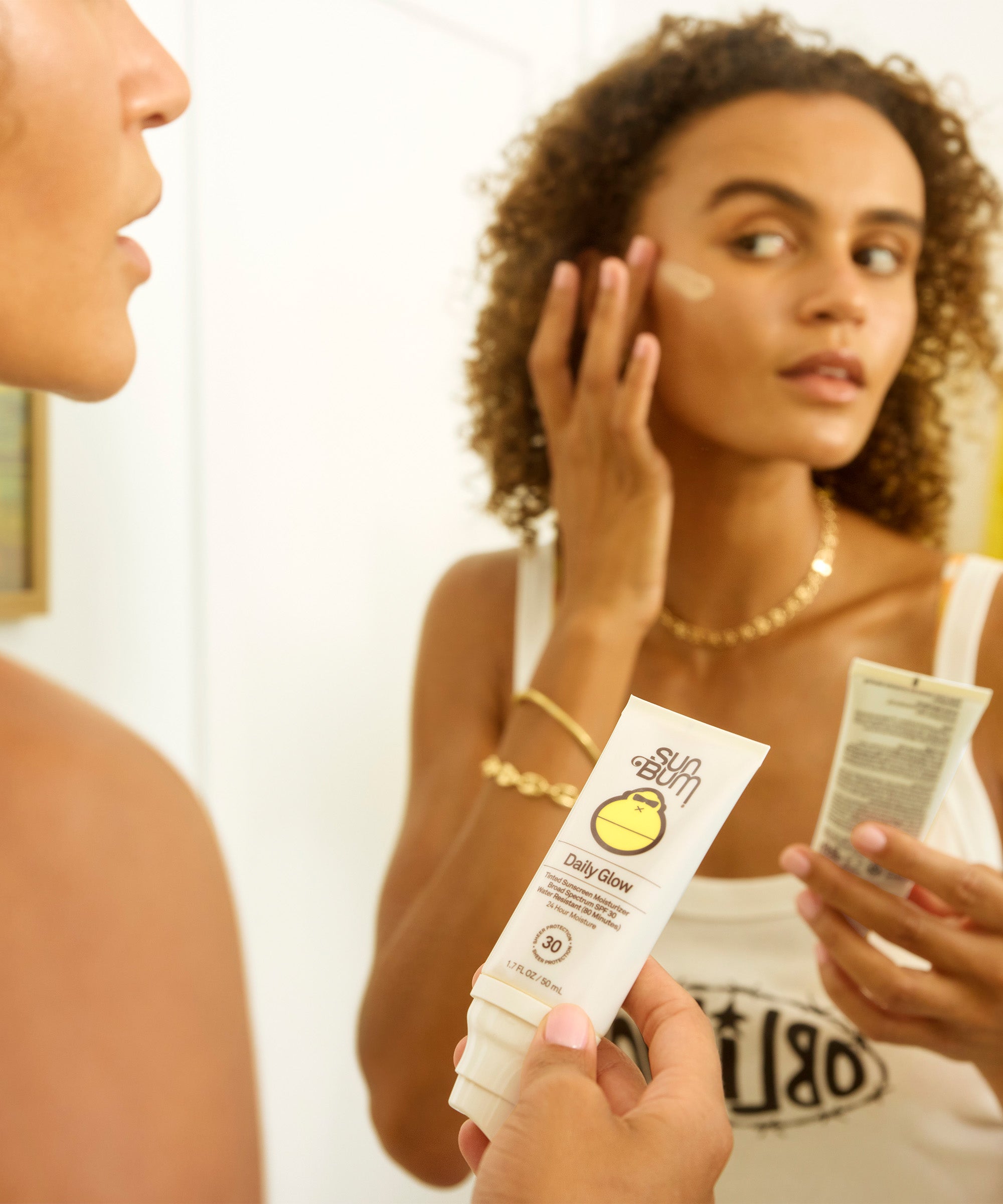 Two women applying Daily 30 Glow moisturizer with SPF 30, showcasing dewy skin and product packaging for all-day hydration and sun protection. Sun Bum