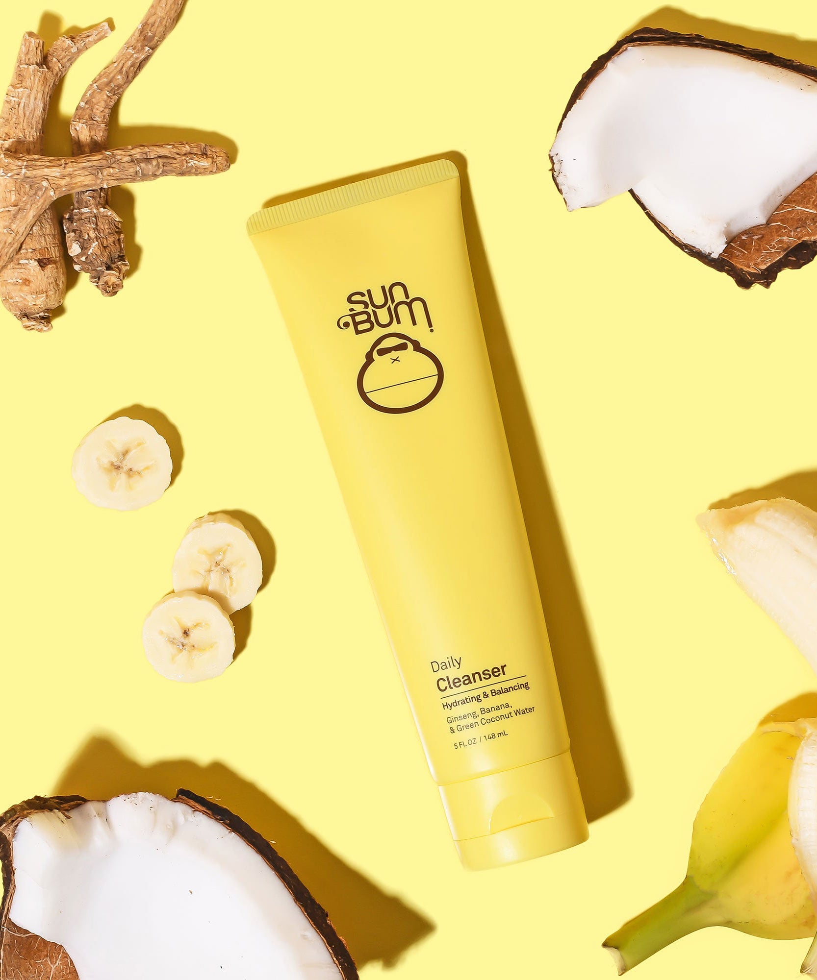 Yellow tube of Daily Cleanser by Sun Bum, surrounded by ginseng roots, coconut pieces, and banana slices on a bright yellow background.