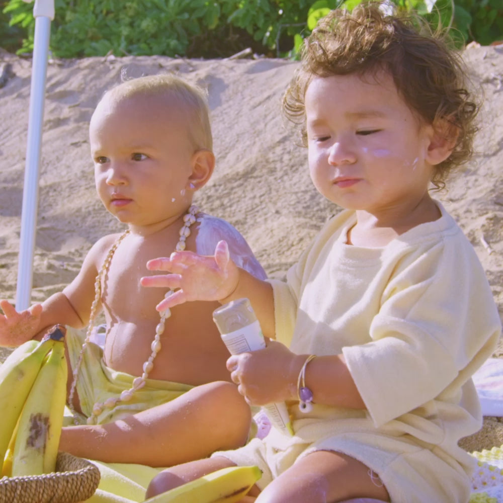 2 babies sitting at the beach holding sunscreen