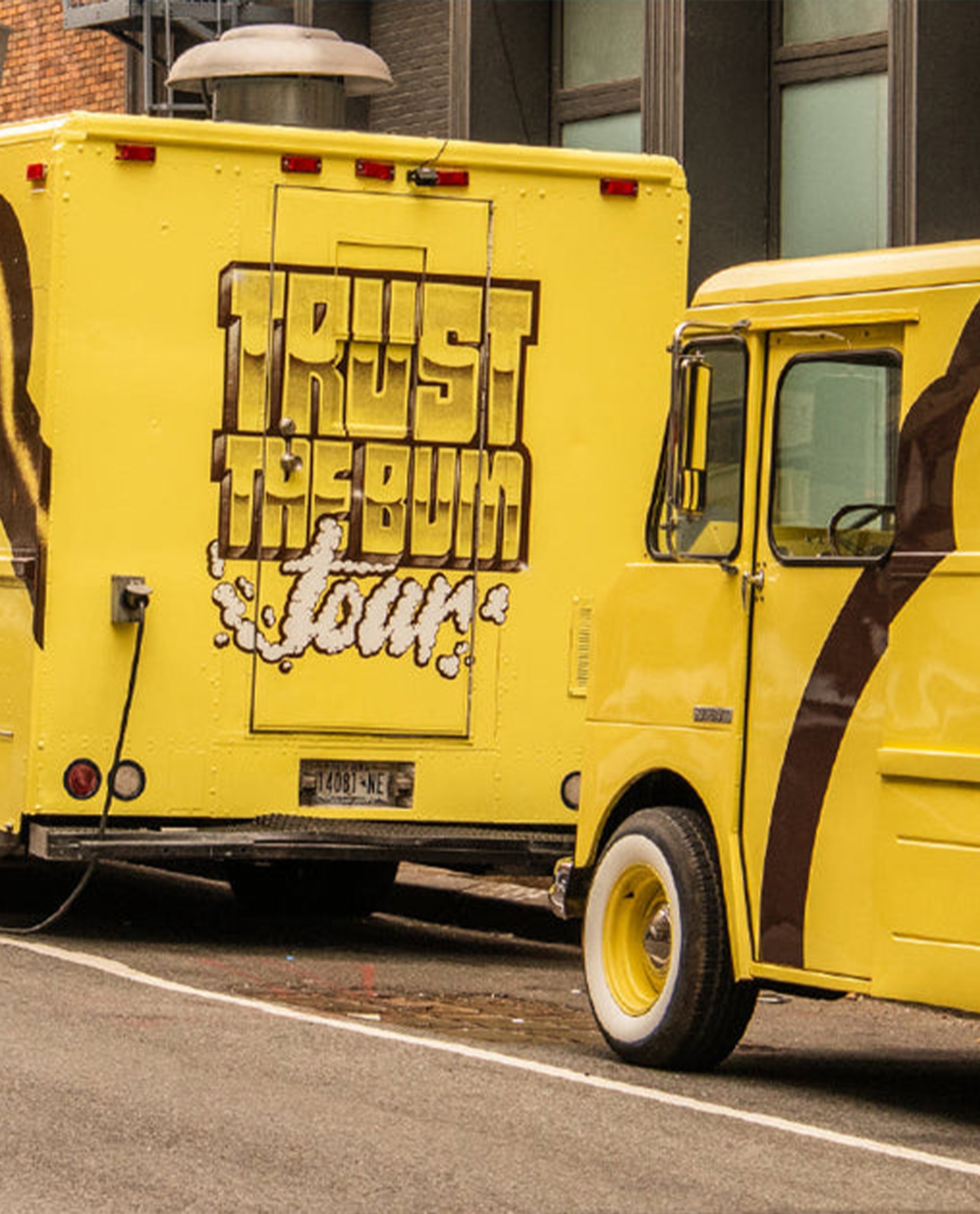 Yellow tour bus with 'Trust the Bum Tour' text on a city street.