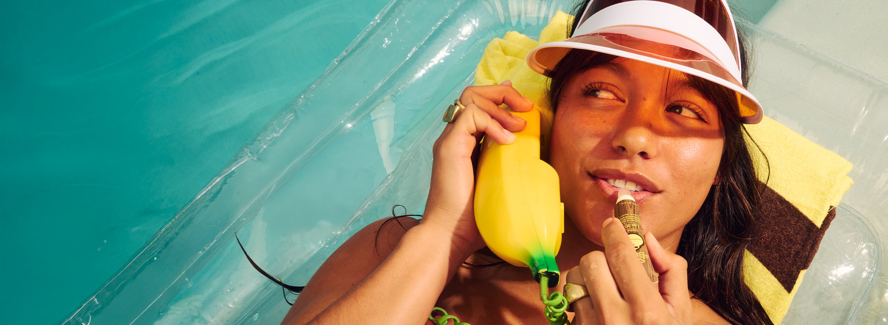Woman lounges poolside on float, chatting on banana phone and applying lip balm – Lip Collection - Sun Bum.