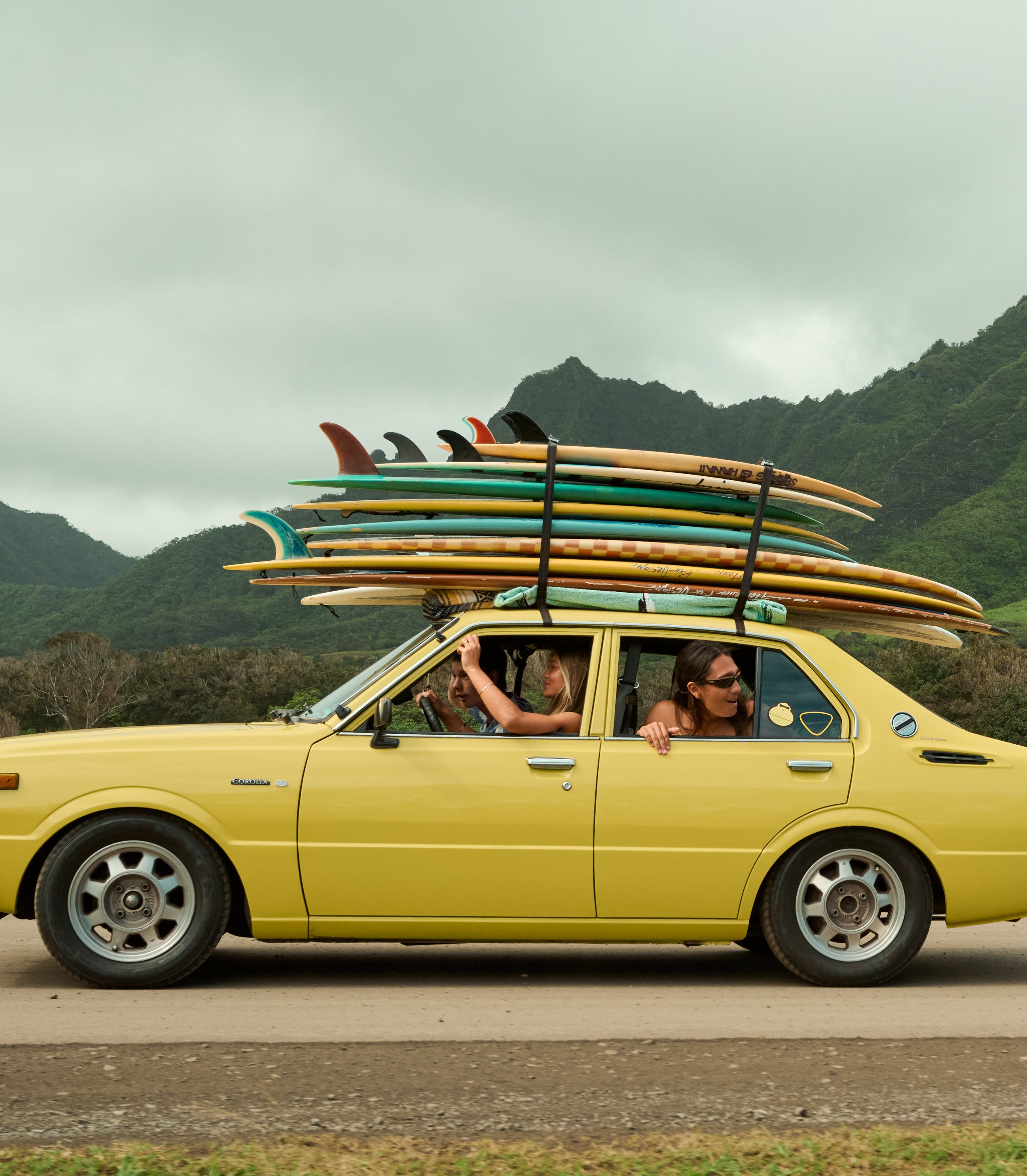 Friends driving a yellow car stacked with surfboards through lush mountains, featuring the SB After Sun Care Collection – Sun Bum.
