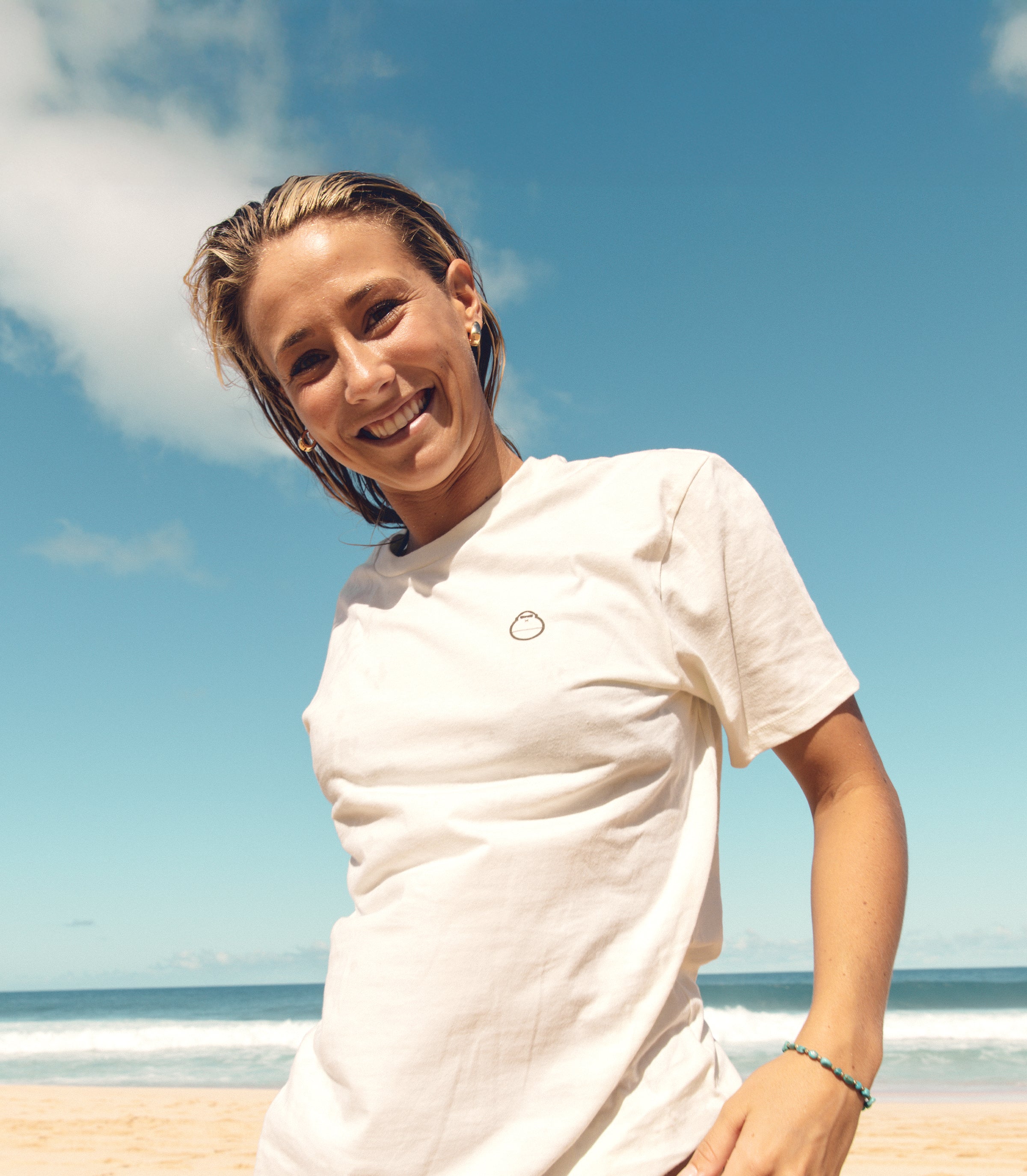 Woman smiling on the beach in a white logo tee under sunny skies, featuring the SB Apparel Collection – Sun Bum.