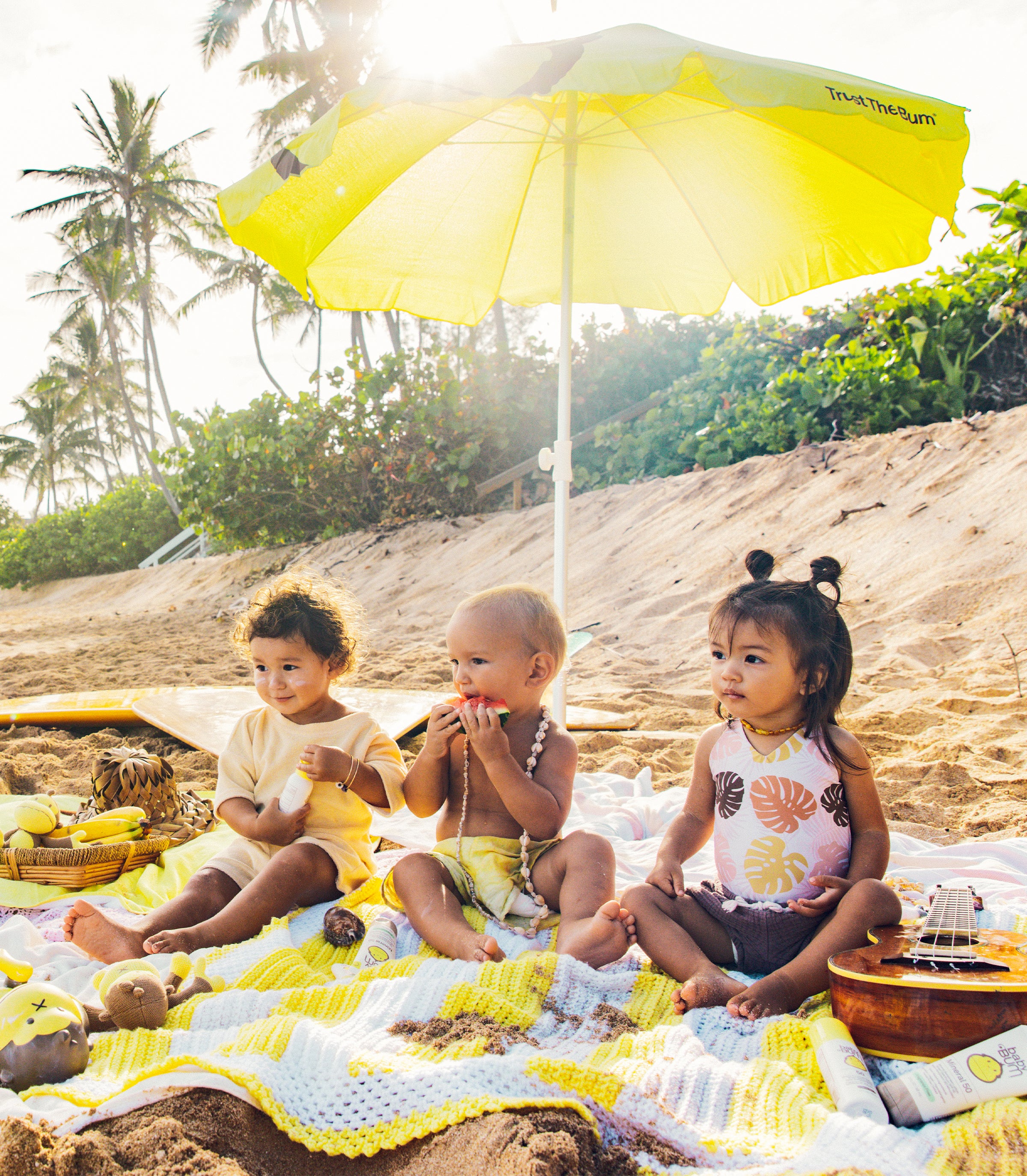 Three toddlers playing on a beach blanket under an umbrella with Baby Bum products from the SB Baby Bum Collection – Sun Bum.
