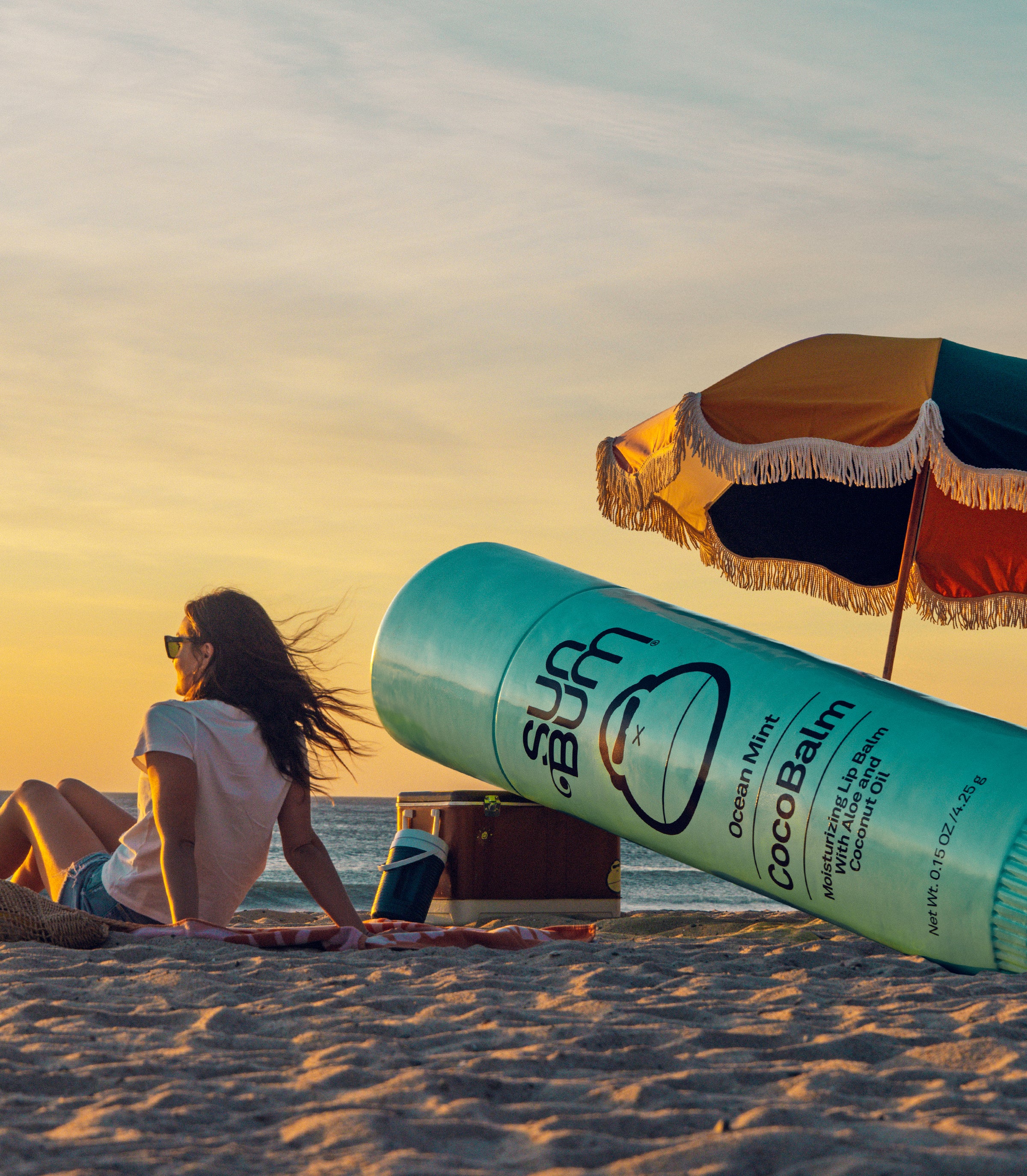 Woman relaxing on the beach at sunset next to oversized CocoBalm lip balm, from the SB CocoBalm Collection – Sun Bum.