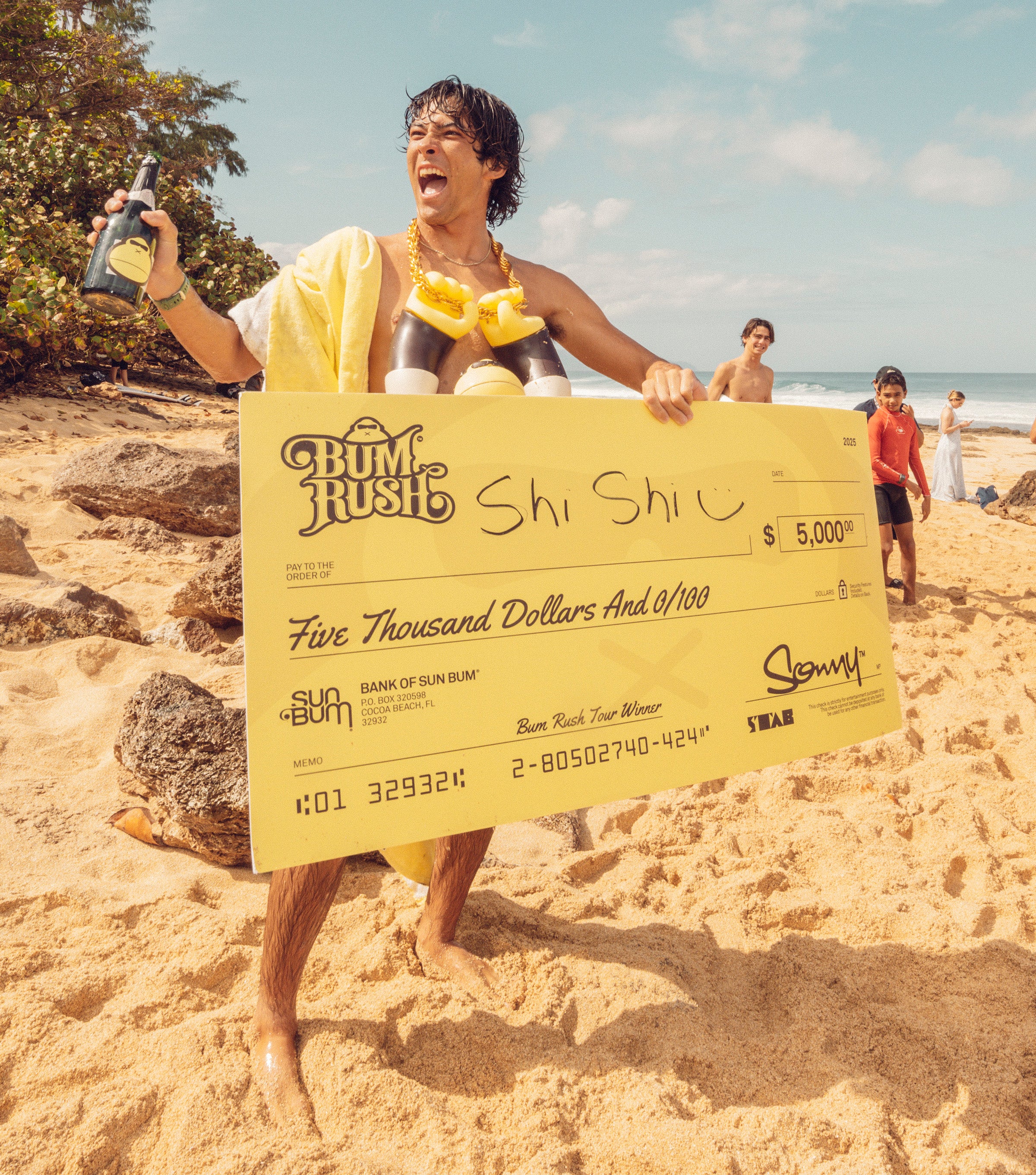 Surfer celebrates on the beach with a giant check and champagne after winning the Bum Rush Tour – Discover Collection - Sun Bum.