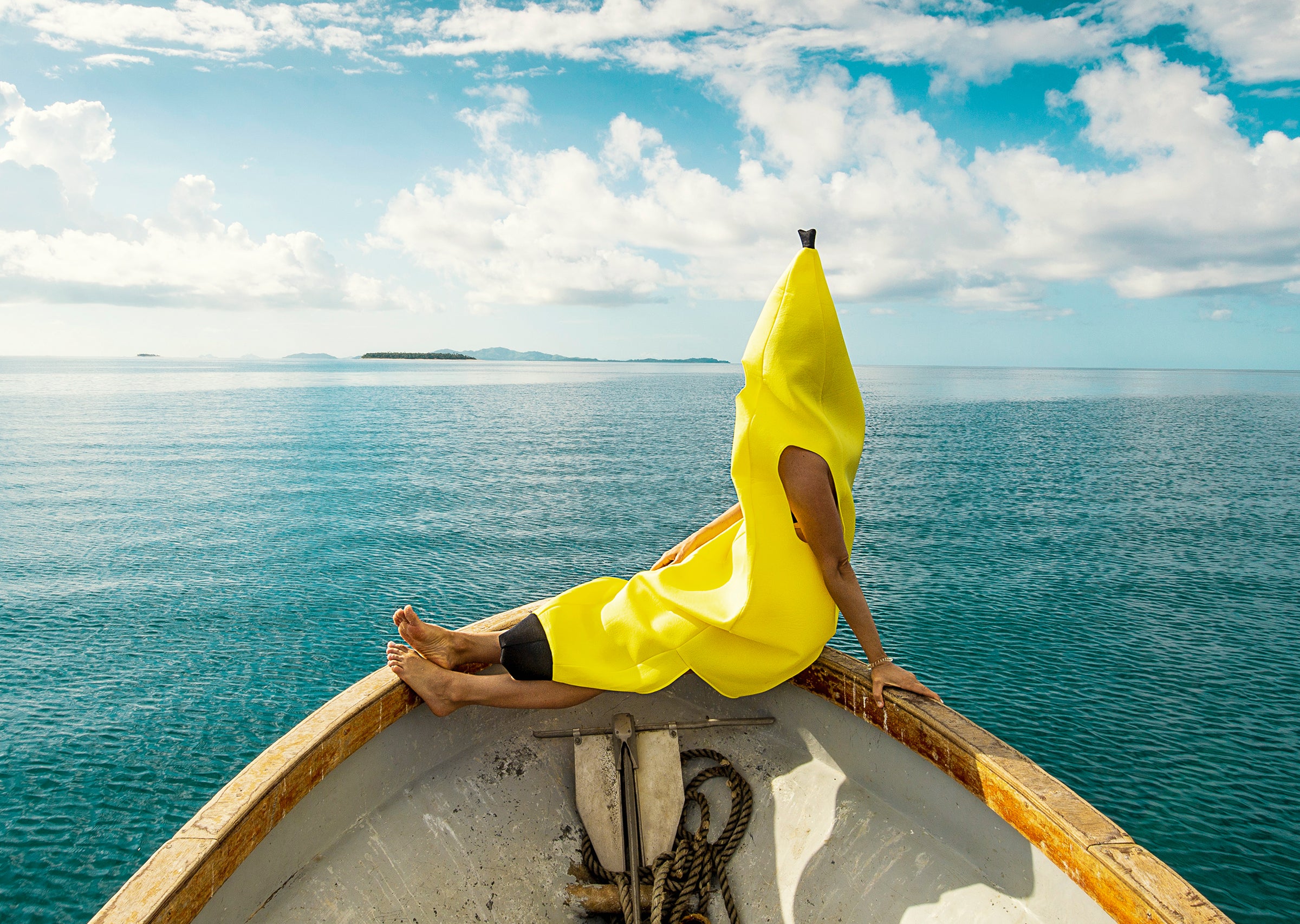 Person in a banana suit relaxing on the bow of a boat in open water, celebrating Banana Suit Fridays Collection - Sun Bum.