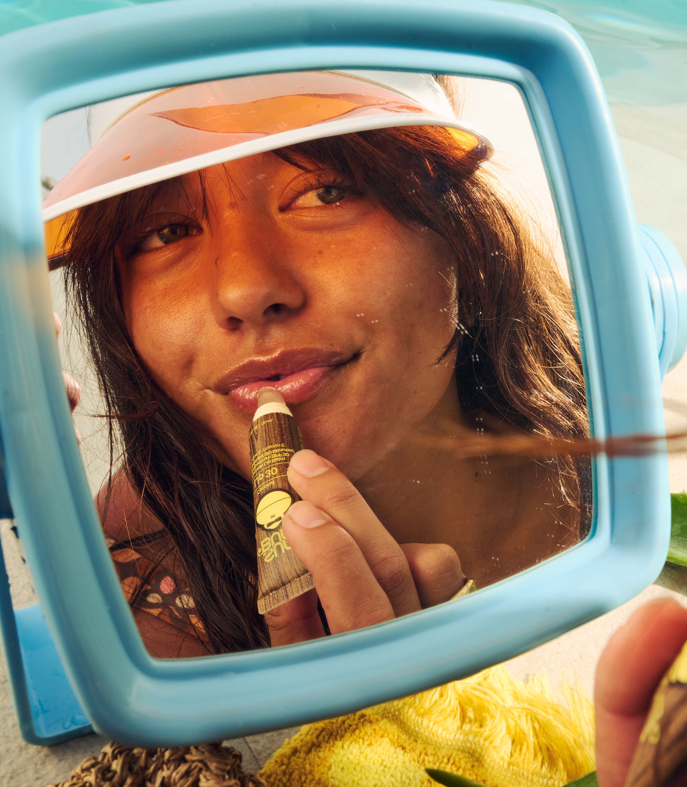 Woman applying SPF lip balm in a mirror reflection while sunbathing, featuring the SB Lip Care Collection – Sun Bum.