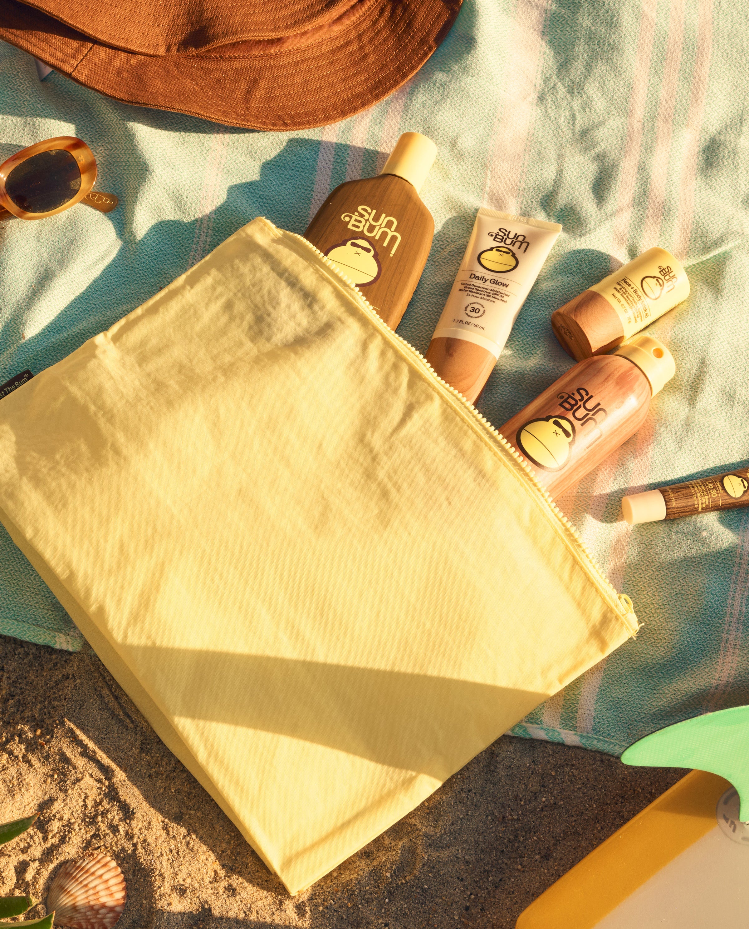 Beige pouch with beach accessories including sunscreen, sunglasses, and a hat on a sandy surface.