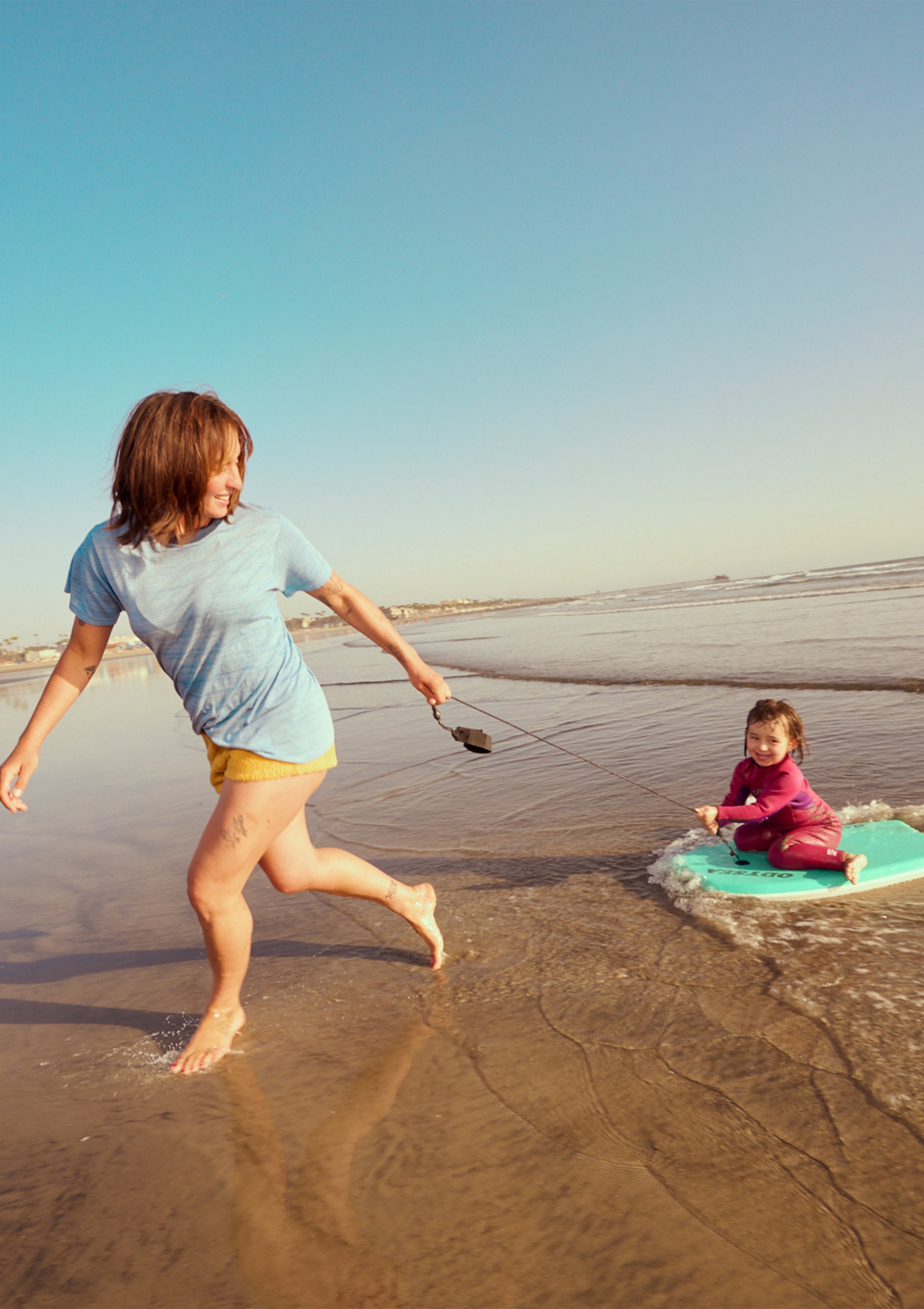 Parent pulling a happy child on a boogie board along the beach at sunset – Promo Blocks Lifestyle Baby Collection - Sun Bum.