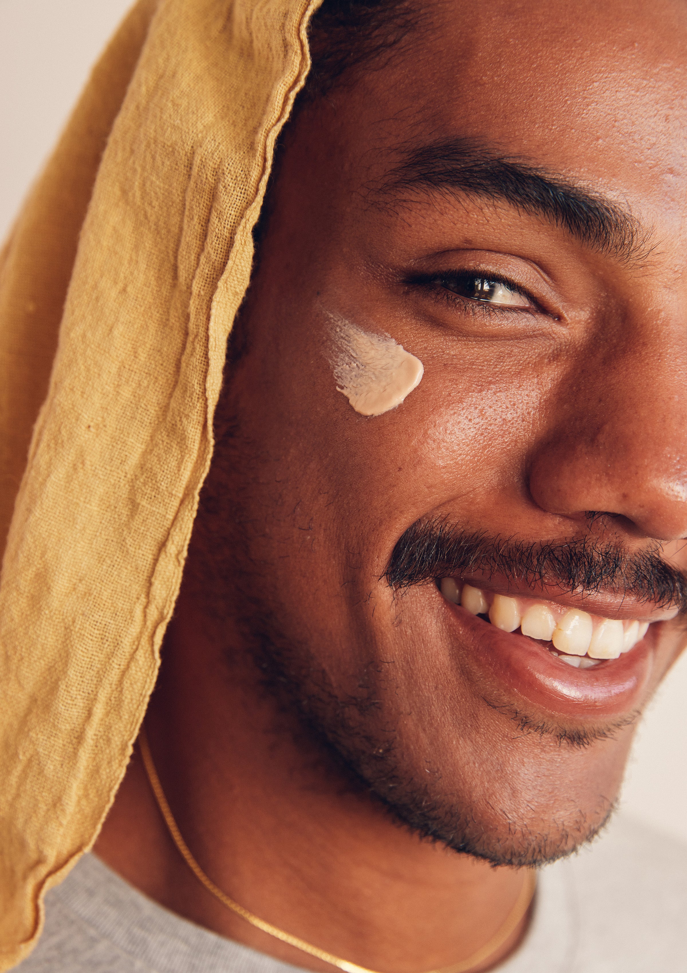 Man smiling with a dab of sunscreen on his cheek, wearing a yellow towel on his head – Promo Blocks Lifestyle Skin Daily Collection - Sun Bum.