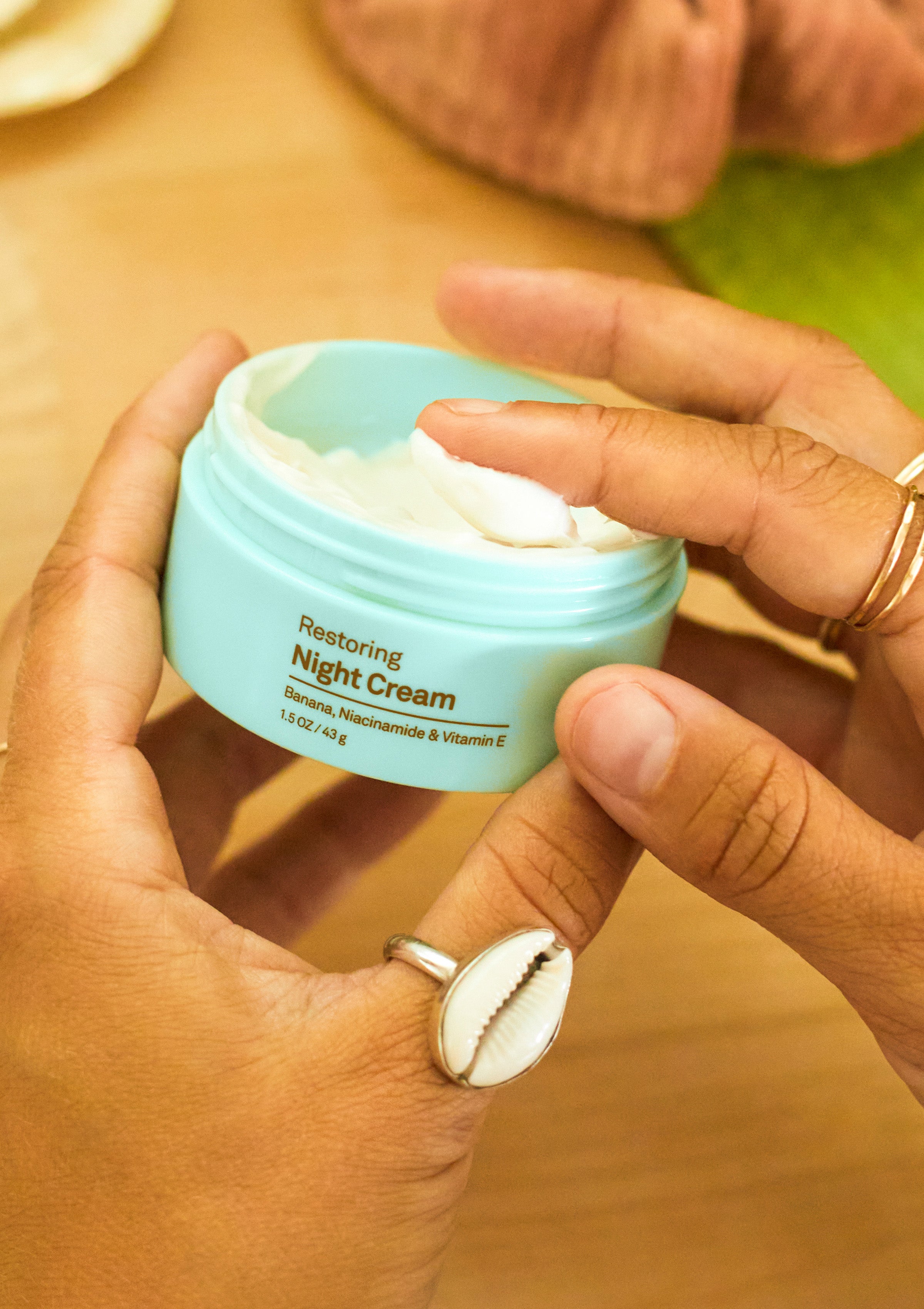 Close-up of hands scooping rich cream from a turquoise jar labeled “Restoring Night Cream” – Promo Blocks Lifestyle Skin Restoring Collection - Sun Bum.