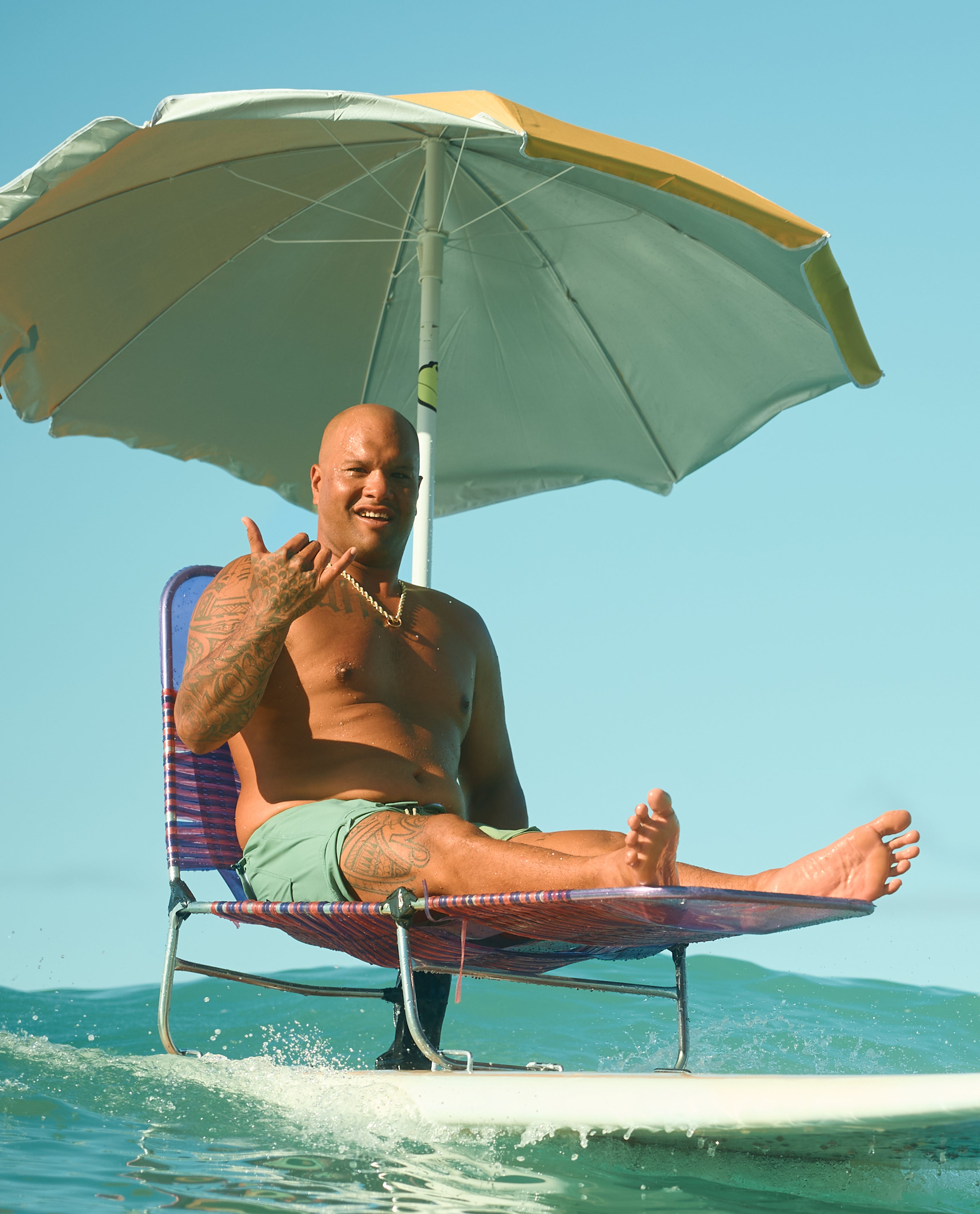 Man lounging on a surfboard chair under a beach umbrella, flashing a shaka in the ocean – Sun Shade Collection - Sun Bum