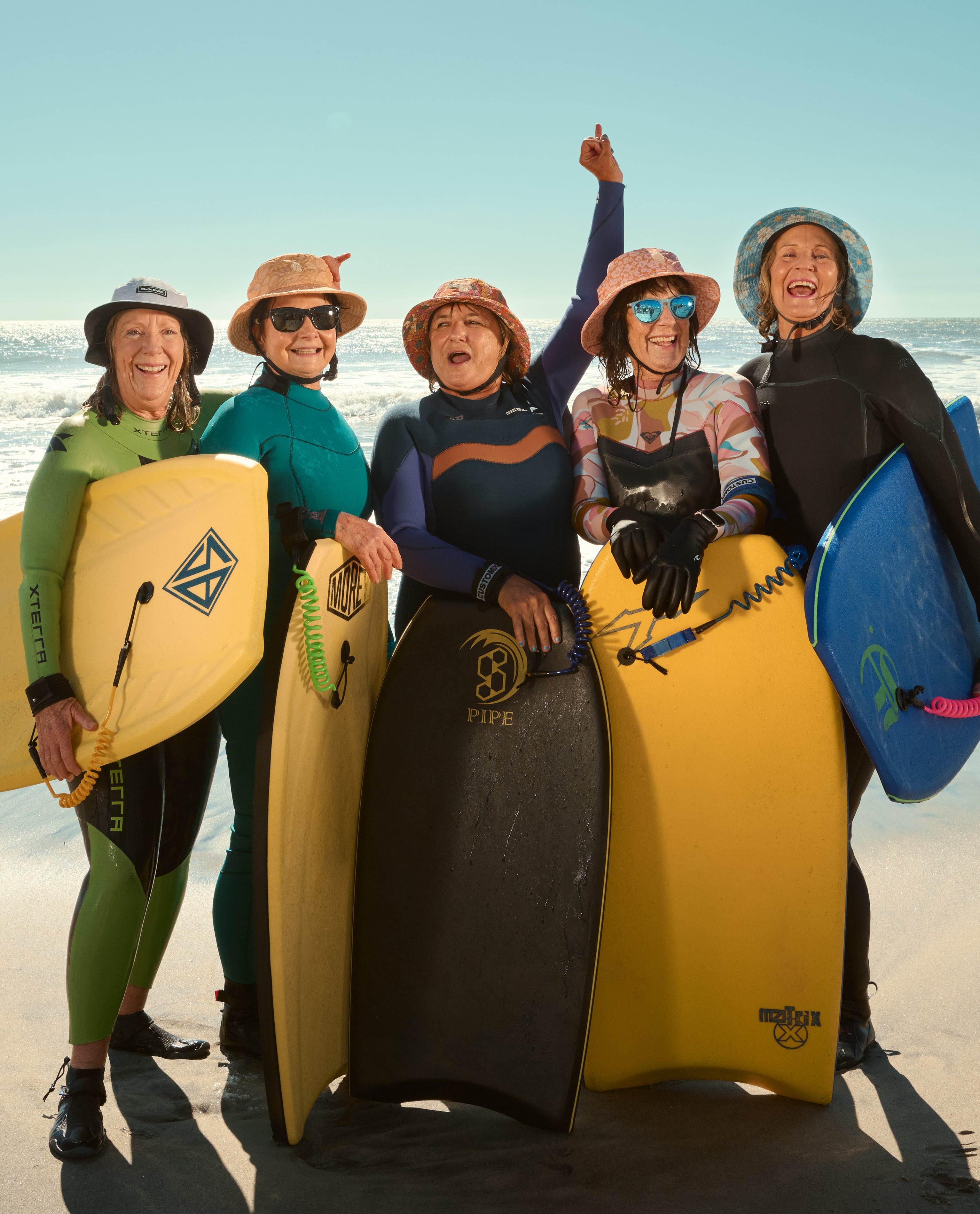 Group of joyful women in wetsuits and hats holding bodyboards on the beach, ready to surf – Sun Sleeves Collection - Sun Bum