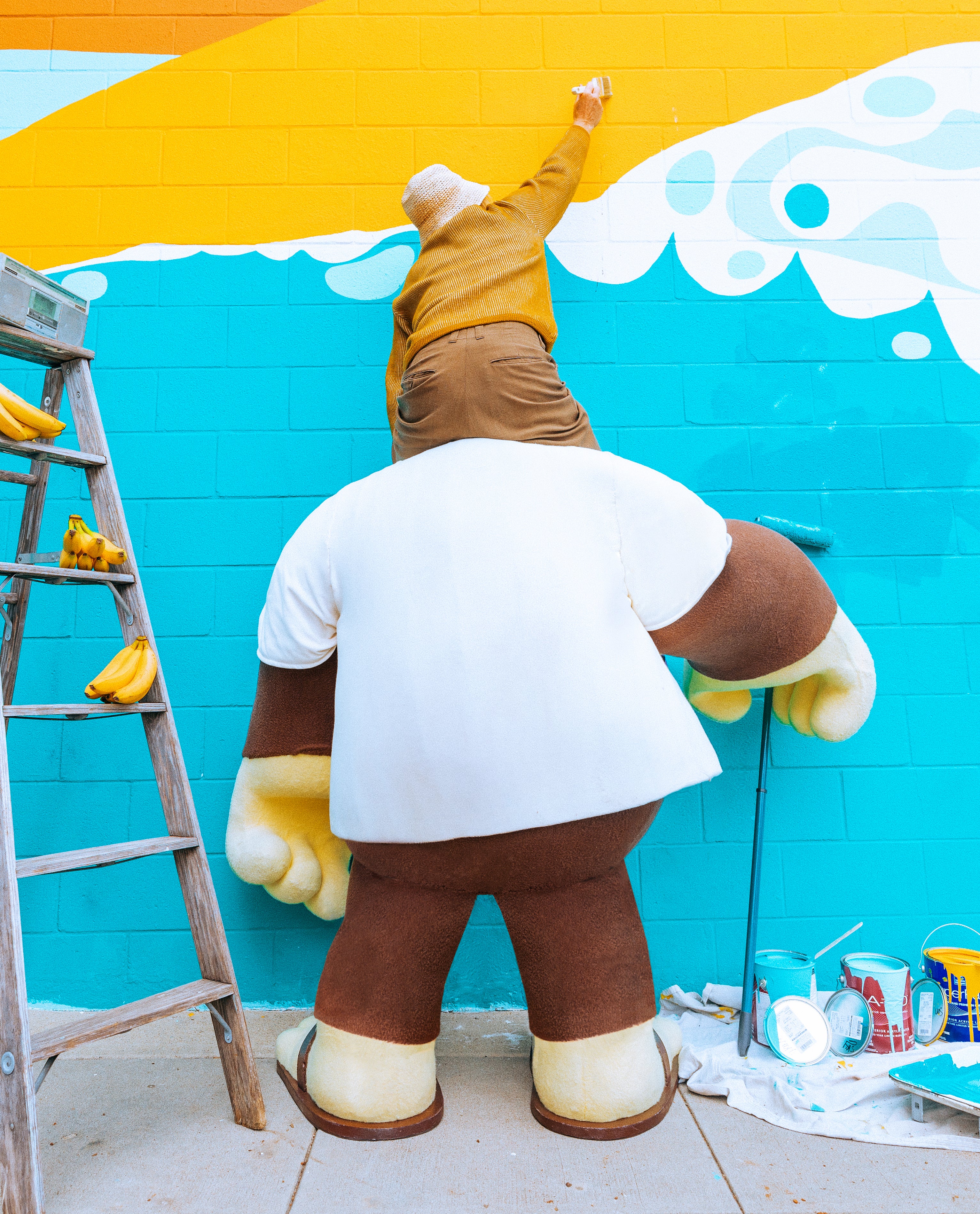 Person painting mural while standing on Sun Bum mascot’s shoulders, surrounded by paint and bananas – Sun Hat Collection - Sun Bum