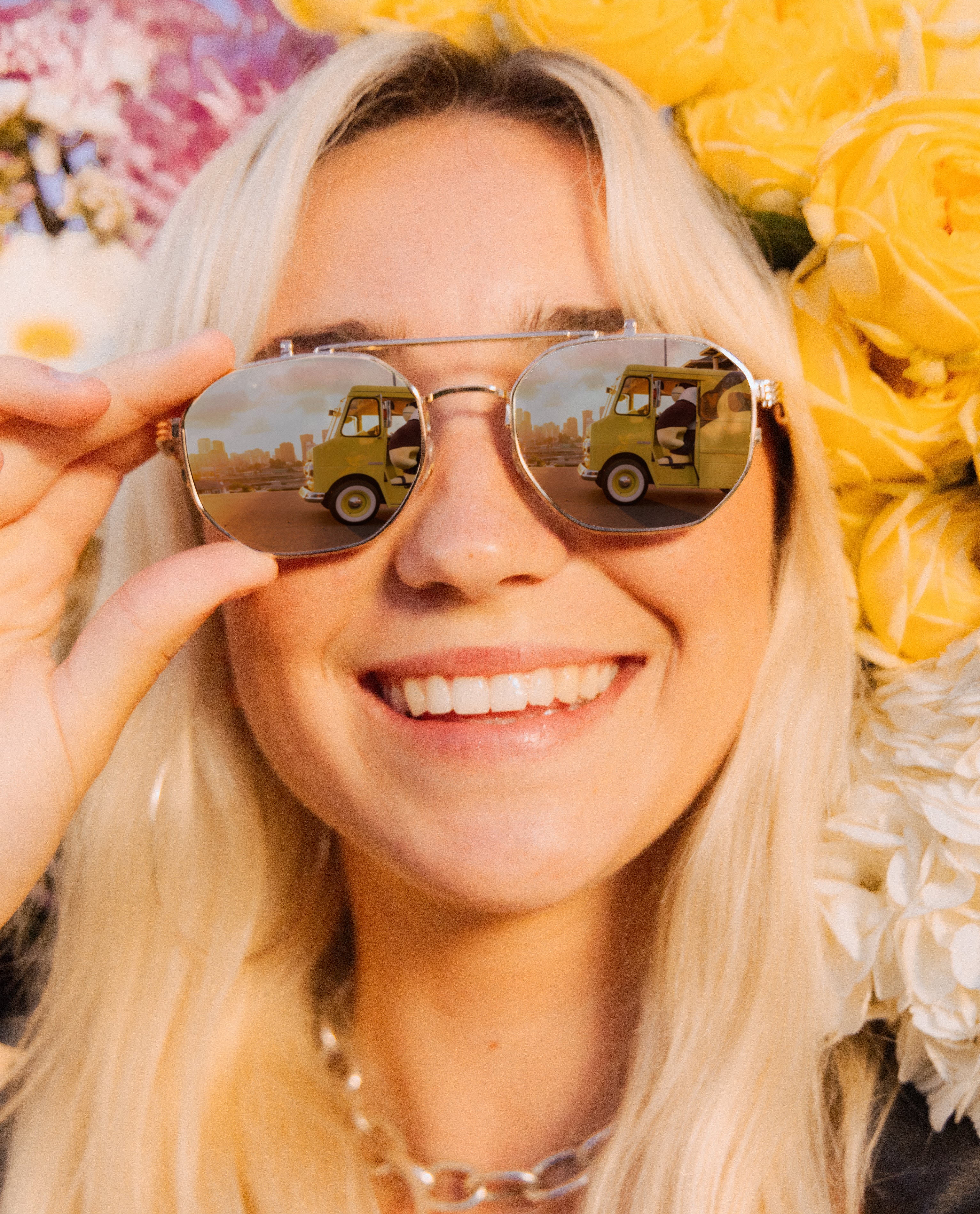 Smiling woman in aviators with a yellow Sun Bum van reflection, surrounded by bright flowers – Sun Sunglasses Collection - Sun Bum