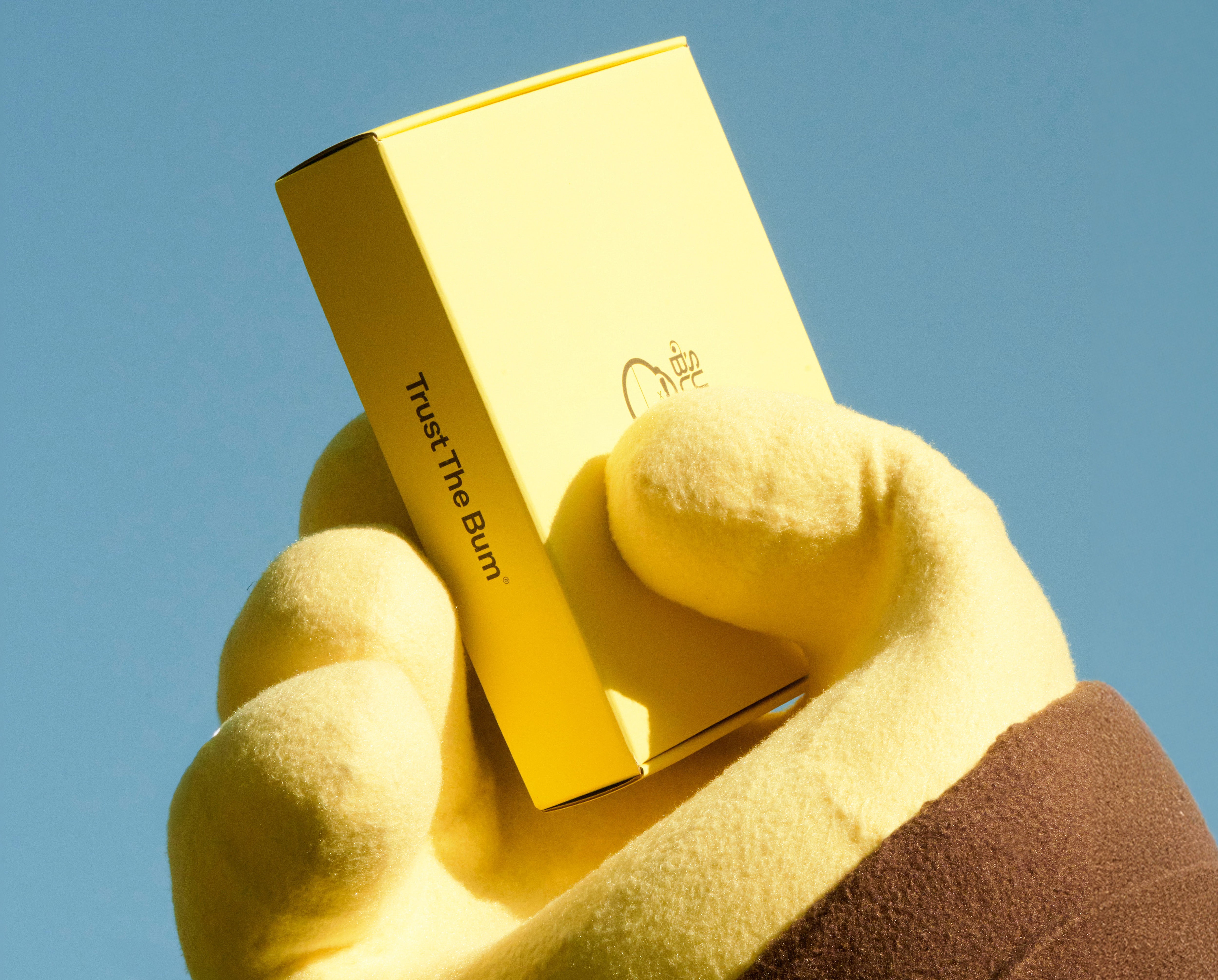 Hand holding a gold product with 'Trust The Bum' text against a clear blue sky