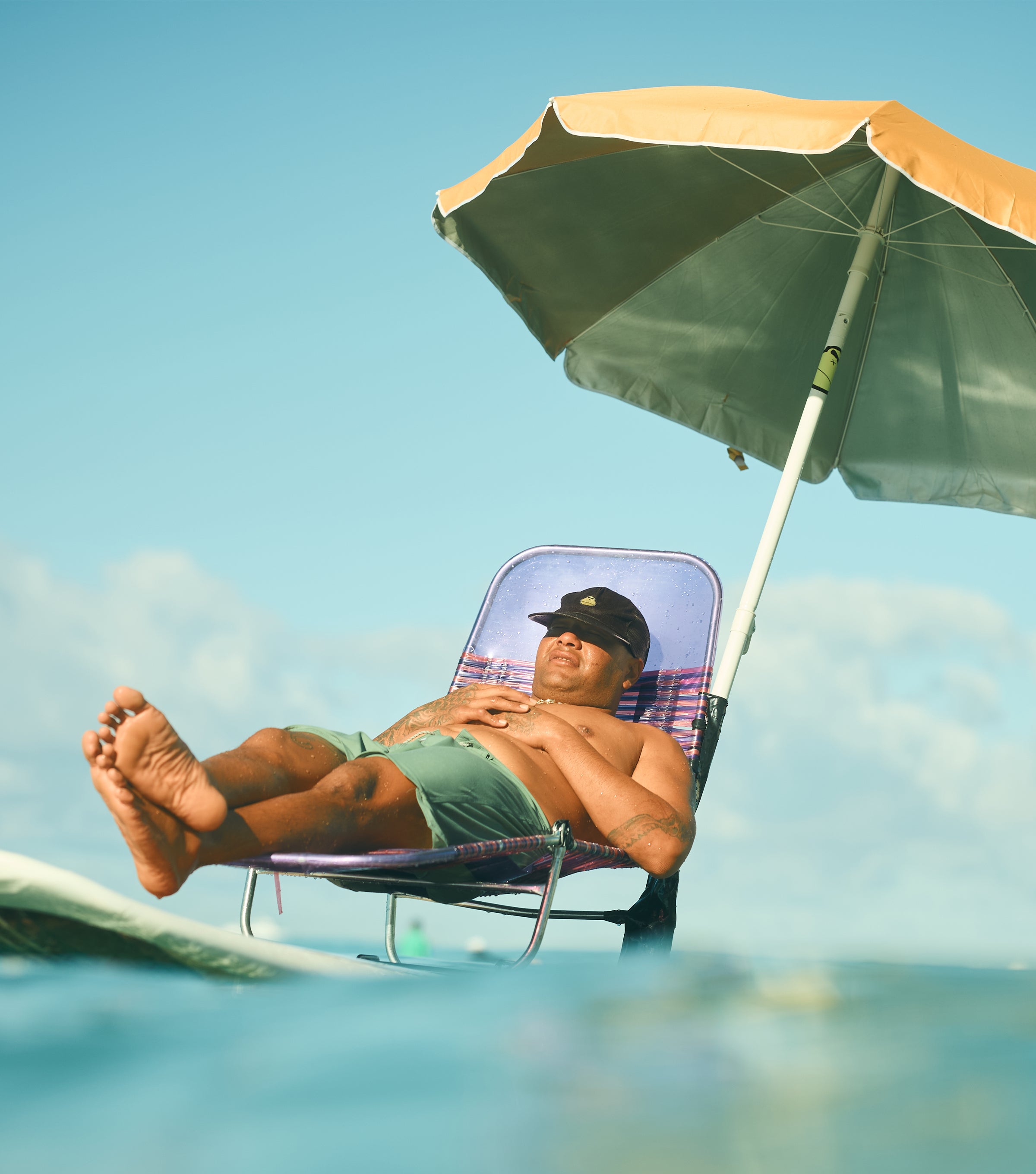 Person lounging on a surfboard in the ocean under a beach umbrella, promoting SEP Sun Education Collection - Sun Bum.