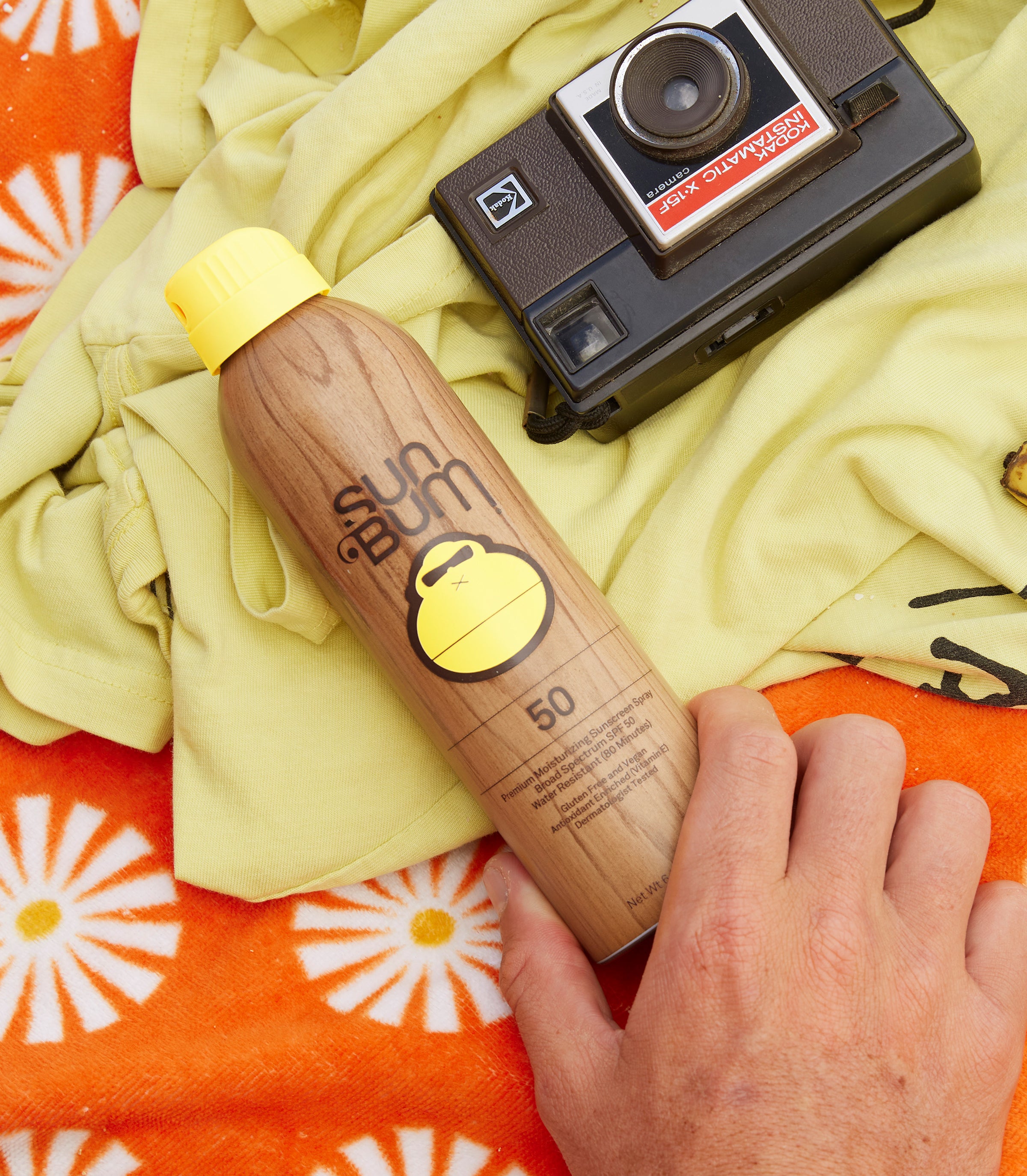 Hand holding Sun Bum SPF 50 spray next to vintage camera on beach towel – Sunscreen Sprays and Mist Collection - Sun Bum.